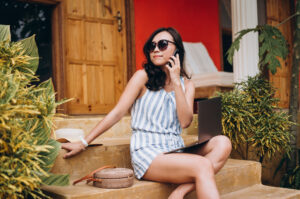 Young Asian woman wearing sunglasses has phone conversation on villa stoop while working from laptop.